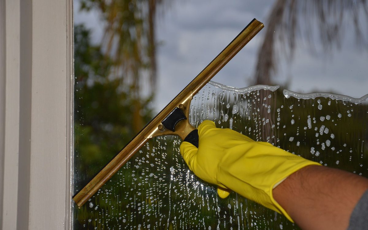 Hand mit gelbem Handschuh zieht eine nasse Fensterscheibe mit einem Abzieher ab